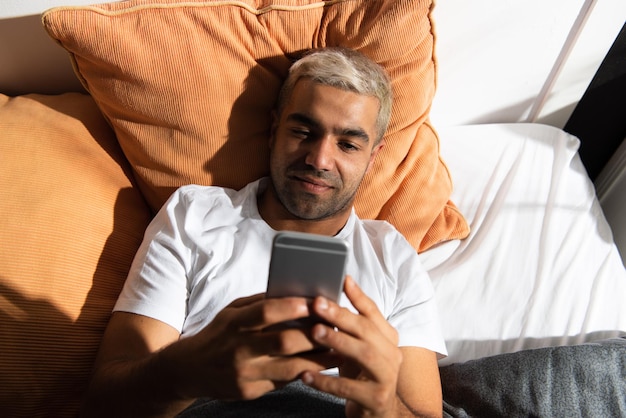 Hispanic man using his phone laying on bed. Bad mornig routine concept.