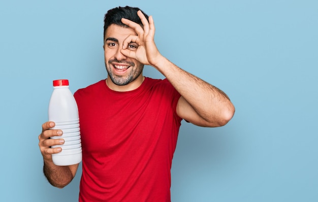 Hispanic man with beard holding liter bottle of milk smiling happy doing ok sign with hand on eye looking through fingers