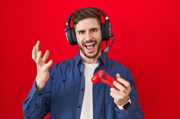 Hispanic man with beard playing video game holding controller celebrating victory with happy smile and winner expression with raised hands