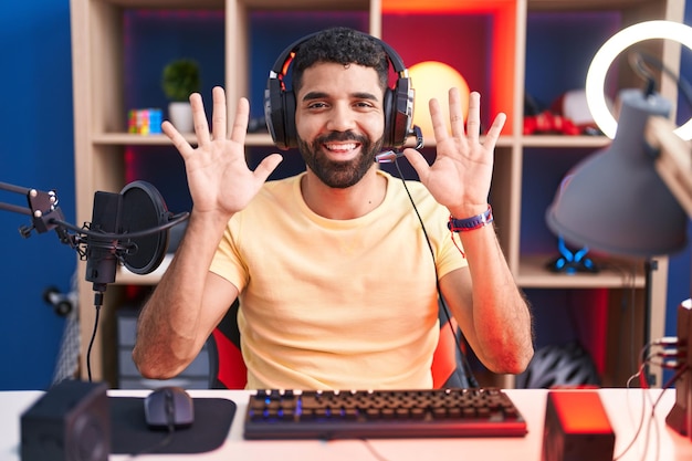 Латиноамериканец с бородой играет в видеоигры в наушниках, показывая и указывая пальцами номер десять, улыбаясь уверенно и счастливо