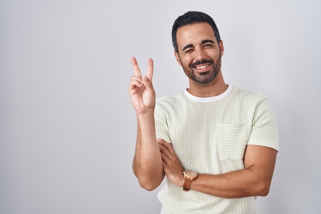 Hispanic man with beard standing over isolated background smiling with happy face winking at the camera doing victory sign with fingers number two