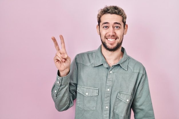 Hispanic man with beard standing over pink background smiling with happy face winking at the camera doing victory sign. number two.