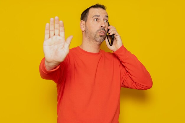 Hispanic man with beard talking on mobile phone pissed off making a stop sign with hand isolated over yellow background person