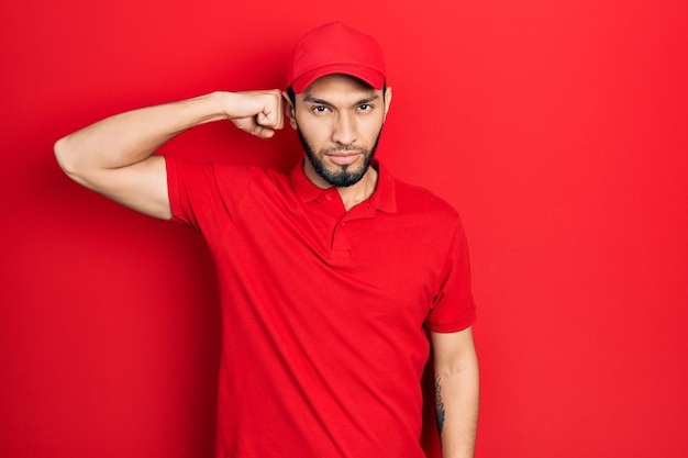 Foto uomo ispanico con barba che indossa uniforme di consegna e berretto persona forte che mostra il muscolo del braccio fiducioso e orgoglioso del potere