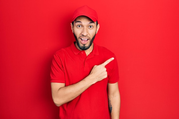 Hispanic man with beard wearing delivery uniform and cap surprised pointing with finger to the side open mouth amazed expression