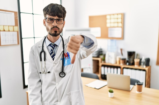 Hispanic man with beard wearing doctor uniform and stethoscope at the office looking unhappy and angry showing rejection and negative with thumbs down gesture bad expression