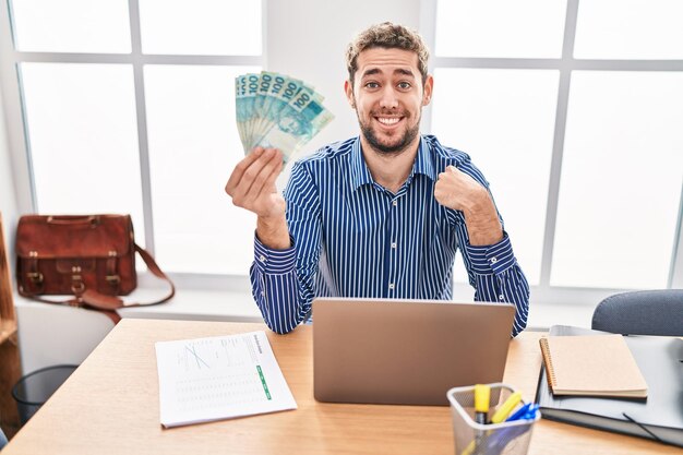 Hispanic man with beard working at the office holding brazilian reals pointing finger to one self smiling happy and proud