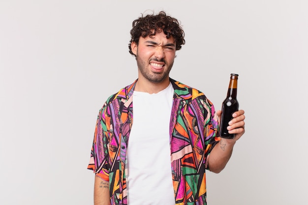 Hispanic man with beer feeling disgusted and irritated, sticking tongue out, disliking something nasty and yucky