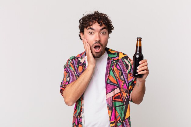 Hispanic man with beer feeling shocked and scared, looking terrified with open mouth and hands on cheeks