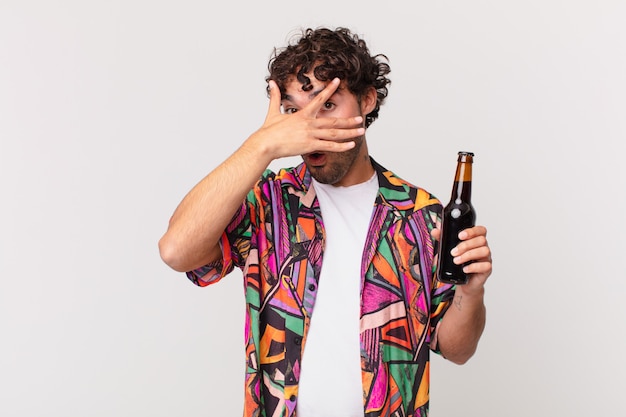 Hispanic man with beer looking shocked, scared or terrified, covering face with hand and peeking between fingers