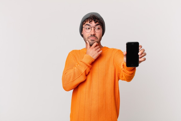 Hispanic man with a cell thinking, feeling doubtful and confused, with different options, wondering which decision to make