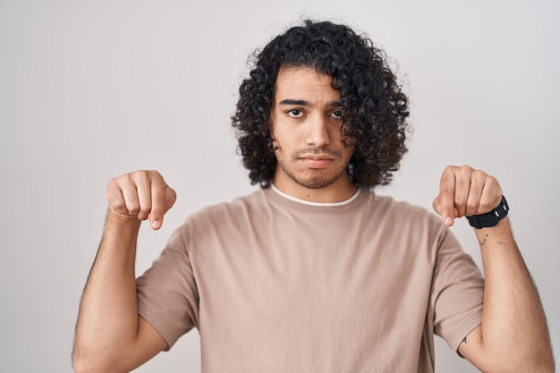 Foto uomo ispanico con capelli ricci in piedi su sfondo bianco rivolto verso il basso guardando triste e sconvolto, indicando la direzione con le dita, infelice e depresso.