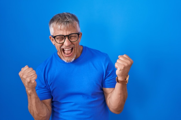 Hispanic man with grey hair standing over blue background very happy and excited doing winner gesture with arms raised, smiling and screaming for success. celebration concept.