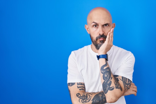 Hispanic man with tattoos standing over blue background thinking looking tired and bored with depression problems with crossed arms.