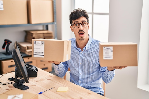 Hispanic man working at small business ecommerce holding packages in shock face looking skeptical and sarcastic surprised with open mouth