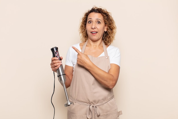 Hispanic middle age woman looking excited and surprised pointing to the side and upwards to copy space