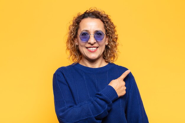 Hispanic middle age woman smiling cheerfully, feeling happy and pointing to the side and upwards, showing object in copy space