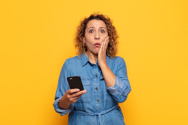 Hispanic Middle aged woman feeling shocked and scared, looking terrified with open mouth and hands on cheeks