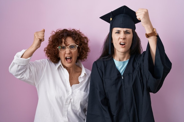 Madre e figlia ispaniche che indossano il cappello di laurea e l'abito da cerimonia arrabbiati e pazzi che alzano il pugno frustrati e furiosi mentre gridano con rabbia. rabbia e concetto aggressivo.