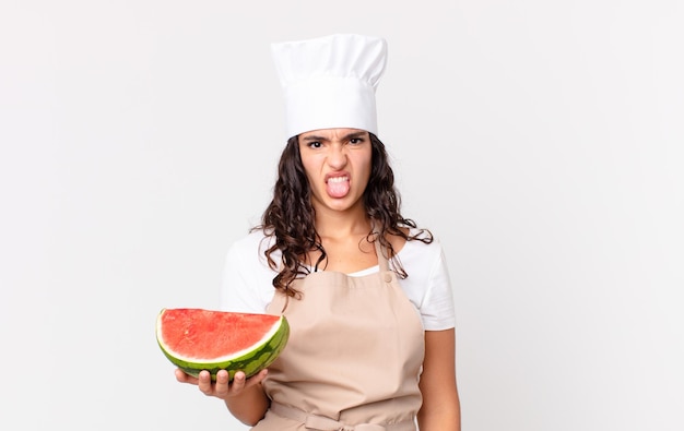Hispanic pretty chef woman feeling disgusted and irritated and tongue out and holding a watermelon