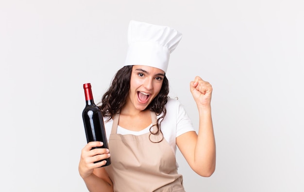 Hispanic pretty chef woman feeling shocked,laughing and celebrating success and holding a wine bottle