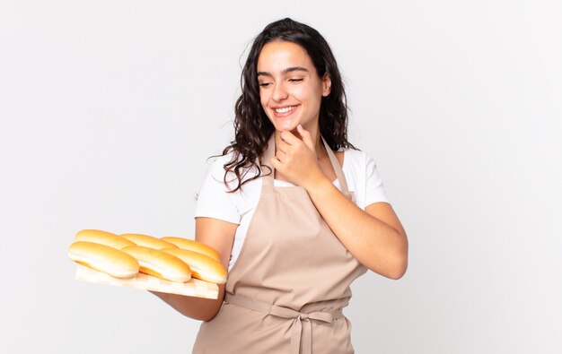 Photo hispanic pretty chef woman smiling with a happy, confident expression with hand on chin and holding a bread buns troy