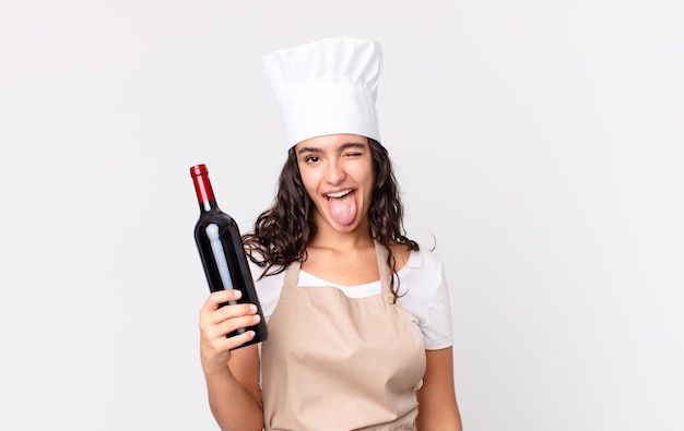 Hispanic pretty chef woman with cheerful and rebellious attitude, joking and sticking tongue out and holding a wine bottle