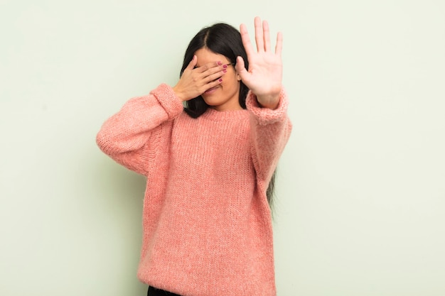 Hispanic pretty woman covering face with hand and putting other\
hand up front to stop camera refusing photos or pictures