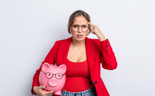 Hispanic pretty woman feeling confused and puzzled, showing you are insane. piggy bank concept