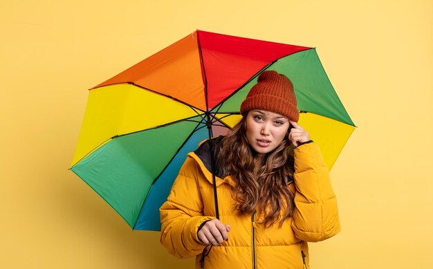 Hispanic pretty woman feeling confused and puzzled, showing you are insane. umbrella concept
