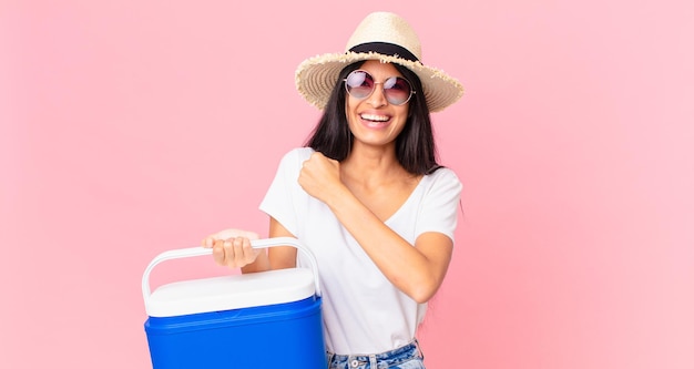 Hispanic pretty woman feeling happy and facing a challenge or celebrating with a picnic portable refrigerator