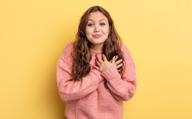 Hispanic pretty woman feeling shocked and surprised, smiling, taking hand to heart, happy to be the one or showing gratitude