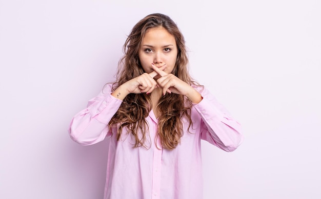 Hispanic pretty woman looking serious and displeased with both fingers crossed up front in rejection, asking for silence