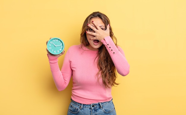 Hispanic pretty woman looking shocked, scared or terrified, covering face with hand. alarm clock concept