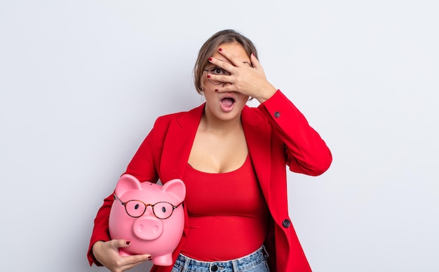 Hispanic pretty woman looking shocked, scared or terrified, covering face with hand. piggy bank concept