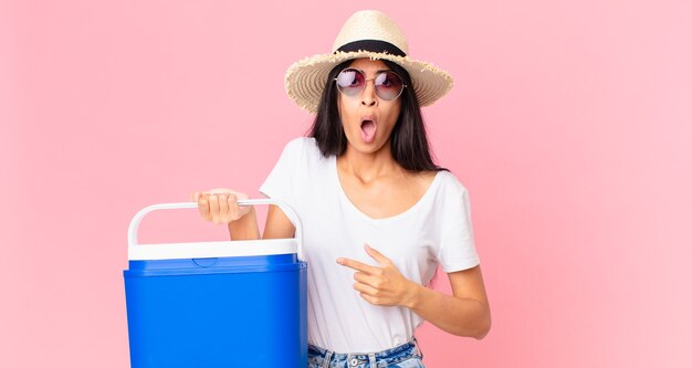 Hispanic pretty woman looking shocked and surprised with mouth wide open, pointing to self with a picnic portable refrigerator