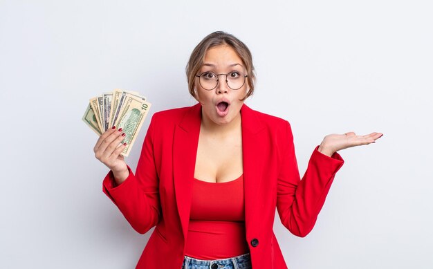 Hispanic pretty woman looking surprised and shocked with jaw dropped holding an object business and banknotes concept