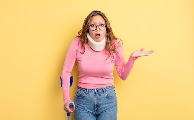 Hispanic pretty woman looking surprised and shocked, with jaw dropped holding an object. crutch and collar accident