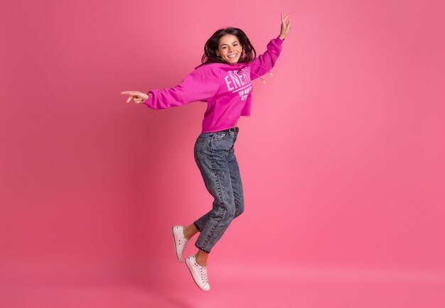 Hispanic pretty woman in pink hoodie and jeans smiling jumping on pink isolated