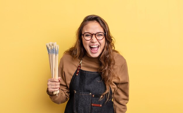 Hispanic pretty woman shouting aggressively looking very angry artist with brushes concept