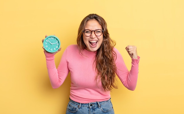 Hispanic pretty woman shouting aggressively with an angry expression. alarm clock concept