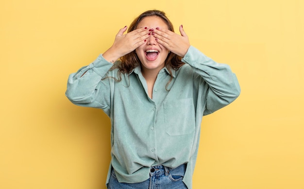 Hispanic pretty woman smiling and feeling happy, covering eyes with both hands and waiting for unbelievable surprise