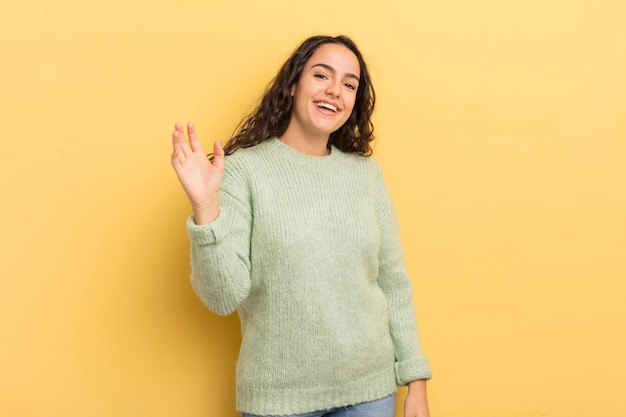 Foto bella donna ispanica che sorride allegramente e allegramente agitando la mano dandoti il benvenuto e salutandoti o salutandoti