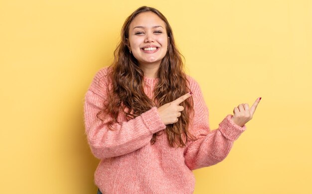 Hispanic pretty woman smiling happily and pointing to side and upwards with both hands showing object in copy space