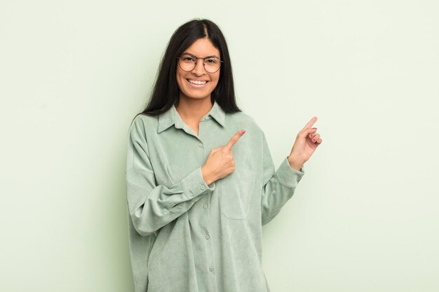 Hispanic pretty woman smiling happily and pointing to side and upwards with both hands showing object in copy space