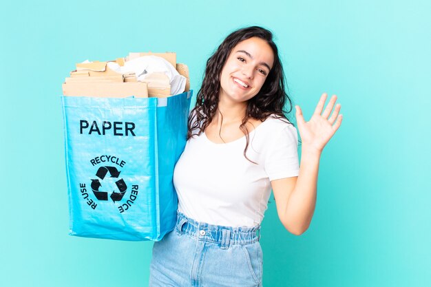 Hispanic pretty woman smiling happily, waving hand, welcoming and greeting you and holding a recycled paper bag