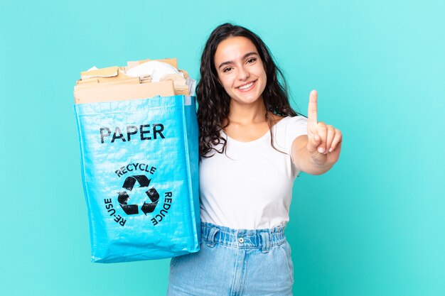 Hispanic pretty woman smiling and looking friendly, showing number one and holding a recycled paper bag