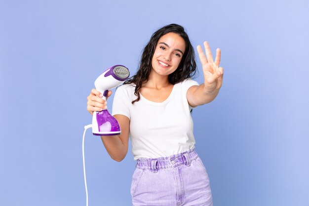 Hispanic pretty woman smiling and looking friendly, showing number three and holding an iron steam