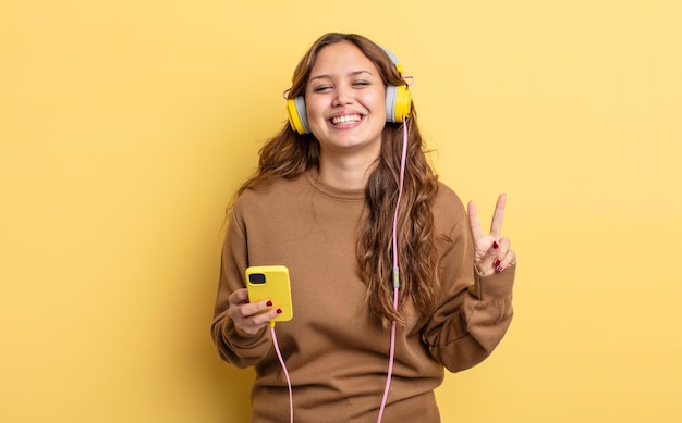 Hispanic pretty woman smiling and looking friendly, showing number two. headphones and smartphone concept