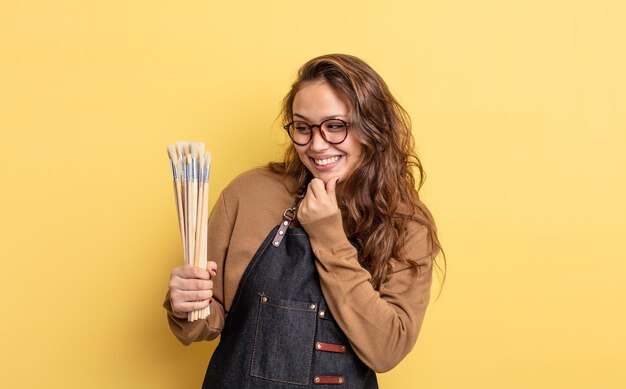 Hispanic pretty woman smiling with a happy confident expression with hand on chin artist with brushes concept